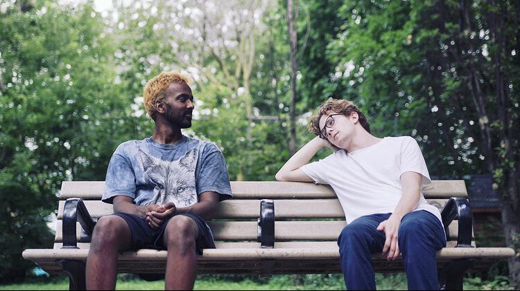 Two actors sit on a park bench in t-shirts. One sits with hands clasped between their knees, while the other leans back, resting their elbow on the back of the bench, and their head in their hand.